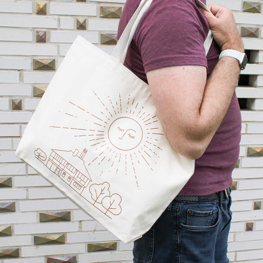This cream canvas tote bag features a line drawing of the Tudor-style Pewabic building with trees around it, in the sky is a huge image of the sun. It has softly closed eyes and a nose with long rays emanating from it.