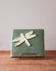 This high relief tile features a dragonfly with detailed wings. It seems to be flying across the tile and sits diagonally, looking to the top right of the tile. The dragonfly is scraped white while the background is a light green color.