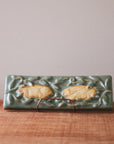 This high relief tile features two young rabbits, resting with ears flat against their backs. They are nestled amongst the green leaves of a protective bush. The rabbits are in scraped of glaze giving them a creamy white color while the rest of the tile is a pale green.