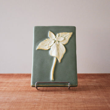This high relief rectangular tile features a large trillium flower. The three petalled flower has a trio of large leaves under it and a slightly curved stem. The flower is scraped to make a creamy white color while the background is a pale green.