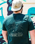 The back of this deep green t-shirt has a large yellow illustration of the Pewabic building surrounded by bushy trees. Above them a large sun with closed eyes and a nose is spreading its rays outward.