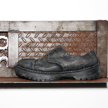 This multimedia sculpture features a life-like ceramic shoe. It is black and worn with missing shoelaces and a thick grippy sole. The work shoe sits on a corrugated metal shelf with decorative gears that like silver metal that is rusting.