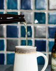 A beer is being poured into a Birch Pewabic stein with a wall of Belle Isle Blend tile behind.