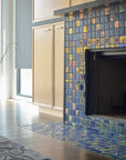 Angled photograph of a blue iridescent tiled fireplace. There is a matte charcoal vase in view. The fireplace is surrounded by light wood cabinets. You can see a small corner of an ornate, gray rug on the floor in front of the fireplace.