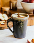 Pewabic Cafe Mug in our matte-black Carbon glaze. The mug is filled with mulled apple cider on a marble countertop. There is a Pewabic bowl in a white Birch glaze filled with red apples in the background.