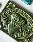 This Claddagh tile features the standard heart, hands and crown design in the center with a Celtic knot motif that wraps above the design and connects the hands- harkening to the original purpose of the Claddagh, as a ring. This tile features the glossy deep green Kale glaze.