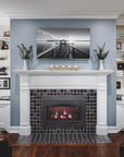 A straight on view of a fireplace and hearth made up of charcoal and glossy black or "onyx" tiles. There is a black and white photo of a dock above the white mantel which holds two "Birch" glazed Pewabic Step Vases. The surrounding white shelves contain personal items such as photos and statues. There are two dark-brown leather couches coming into the corner of the photo.