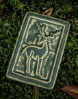 Our 4" x 6" Deer Tile in a muted green "Bayleaf" glaze. You can see the outline of the deer with antlers, which is contrasted a pale-yellow tone. It is laying on a bed of small evergreens surrounded by crisp, brown, autumn leaves.