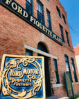 The ceramic Ford Piquette Tile is held up next to the historic Piquette Plant it is designed after.