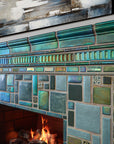 Vibrant, lit fireplace made up of Iridescent and matte glazes which creates an overall green, mirror-like effect. This detailed shot shows the complexity of the custom-cut accent trim tiles.