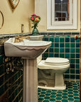 A wooden door opens to a custom tiled bathroom in varying greens and blues. The walls are an off-white color and the bathroom accents colors are mostly white and brass.