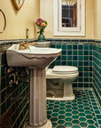 A wooden door opens to a custom tiled bathroom in varying greens and blues. The walls are an off-white color and the bathroom accents colors are mostly white and brass.