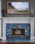 This fireplace features Iridescent tile accents along with a mix of complementary blue and charcoal tones. There are three black candlesticks and a charcoal glazed Pewabic vase on the Colonial style, white mantel. Above the mantel there is a painting of a seaside town.