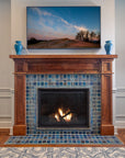 A lively mix of blue, gray, and charcoal tiles are used in this fireplace and hearth set in a Craftsman style wooden mantel. There are two Pewabic Classic Vases on either side of the mantel in our true blue "Peacock" glaze. The fire is lit and there is a landscape photo framed on the wall of a sand dune with wispy trees in the background.