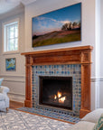 A lively mix of blue, gray, and charcoal tiles are used in this fireplace and hearth set in a Craftsman style wooden mantel. The fire is lit and there is a landscape photo framed on the wall of a sand dune with wispy trees in the background.