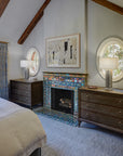 A neo-futuristic fireplace and hearth comprised of Iridescent tiles which are green/blue and mirror-like. There is a framed piece of abstract art above the fireplace with wooden dressers, round windows, and lamps on either side.