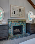 A neo-futuristic fireplace and hearth comprised of Iridescent tiles which are green/blue and mirror-like. There is a framed piece of abstract art above the fireplace with wooden dressers, round windows, and lamps on either side.