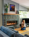 Angled view of a custom Pewabic fireplace set inside of a Midcentury-style home. There is a person holding a small dog near the lit fireplace. The windows on either side of the fireplace bring out the vibrant shine of the tilework.