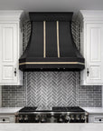 Light Gray Modern Herringbone pattern tiles line the wall behind a stainless steel stove with a large matte black hood. Ornate white cabinets flank the tile on either side.