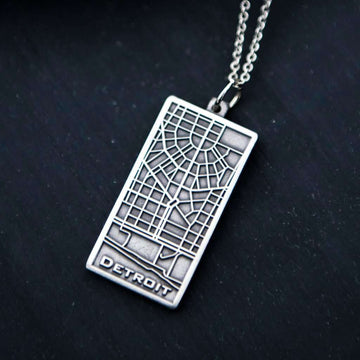This brushed metal Detroit Map Pendant is rectangular with a line drawing of the areal view of a downtown Detroit street map. The word "Detroit" is written at the bottom of the design.