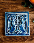 The square tile shows two angels in flowing robes with large feathered wings face each other, each has their hands grasping a bare tree growing between them. This tile features the glossy deep blue Ocean glaze.