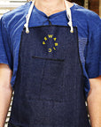 The Pewabic Apron is a dark denim wash with the Pewabic logo in bright yellow in the center of the chest.