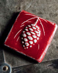 This pinecone tile features the deep glossy red Winterberry glaze. The glaze breaks at the raised portions of the design, letting the ivory of the clay body peek through.