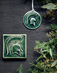 The MSU Tile and Ornament are sitting next to each other. Both pieces feature the same Spartan helmet design.