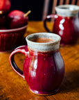 Two mugs sit on a warn wooden table. A Winterberry bowl sits behind them holding red pears.