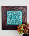 The Lovebirds Tile has a high relief design showing two birds facing one another on a tree branch while pecking at the same berry. There is an ornate border design around it including leaves and twirled lines. The tile features the matte turquoise Pewabic Blue glaze which beautifully offsets the deep reddish brown hues of the oak wood frame.