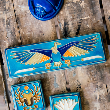 The short and wide Nekhbet tile features a line drawing of a vulture with wings outstretched, head turned to the right as she holds two stylized feathers- one in each talon. This tile features bright shades of blue, orange, cream and white.