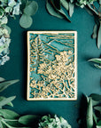 This Mountain View Tile features a a large flowery bush in the foreground with a tall pine tree behind it. In the distance, craggy mountains sit under a cloudy sky.  