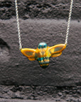 The bee necklace is held up against a black painted brick wall.