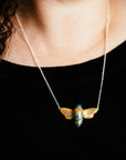 The Bee Necklace features a high-relief bee with wings outstretched. The hand-painted iridescent glaze gives it black stripes and a black head while the rest of the bee features the gold Blush Iridescent glaze. The bee is smooth and shiny and its wings are attached to the silver chain.