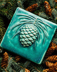 The 6x6 Pinecone tile features a large pinecone in the center of the tile with pine needles encircling it. This piece shown in Pewabic blue- a turquoise blue color.