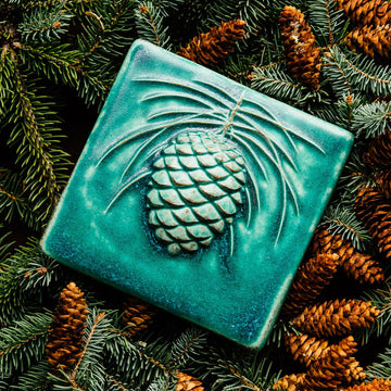 The 6x6 Pinecone tile features a large pinecone in the center of the tile with pine needles encircling it. This piece shown in Pewabic blue- a turquoise blue color.