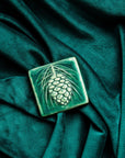 The ceramic Pinecone Tile has a high relief design with one large pinecone in the center and long spindly pine needles framing it. This tile features the matte blueish-green Pewabic Green glaze.