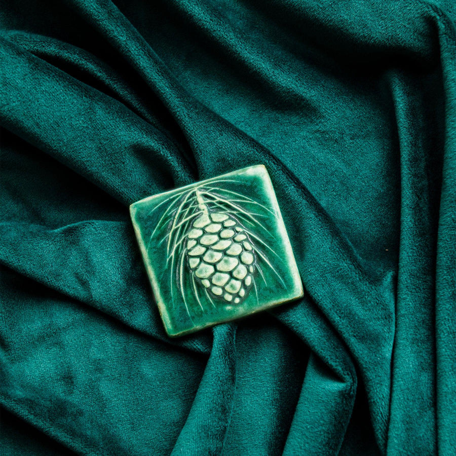 The ceramic Pinecone Tile has a high relief design with one large pinecone in the center and long spindly pine needles framing it. This tile features the matte blueish-green Pewabic Green glaze.