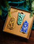 Three ceramic ornaments are sitting in a gift box and placed under a Christmas tree.