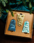 Three ceramic ornaments are sitting in a gift box and placed under a Christmas tree.