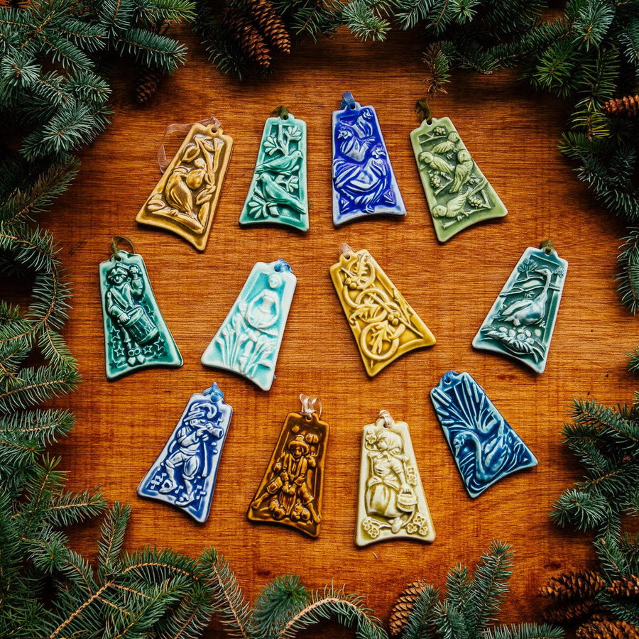 A full set of 12 Days of Christmas ceramic ornaments are haphazardly laid on a wooden surface with boughs of greenery surrounding them.