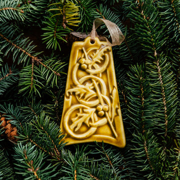 The bell-shaped Five Gold Rings Ornament features five intertwined smooth rings. There are springs of holly leaves and berries throughout the design. The ornament is glazed in a matte yellow glaze.