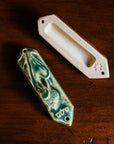 This ceramic Mezuzah features an acorn and oak leaf with the word "Peace" written in Hebrew below them. This piece is thick with an opening on the back to hold your written prayer. There are two holes at the top and bottom to make it easy to attach to your doorframe. This photo features the back of our Mezuzah in a “Leaf” glaze. 