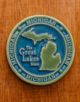 This round Michigan Trivet Tile features the outline of the state's two peninsulas including some of the larger northern islands of Michigan. To the left of the design reads "The Great Lakes State" and surrounding it, in a border, reads the word Michigan four times. This two tone Michigan tile features a matte blue background while the peninsulas and words are in a matte organic green color.