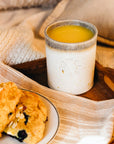 These Rocks cups feature the creamy white Birch glaze that has some brown speckling. The inner lining and lip of the cup have a mottled gray color. A Rocks cup filled with orange juice sits next to a plate covered in scones.