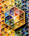 The Hex Paperweight is a thick ceramic tile shaped like a hexagon. The surface of the tile features an interlocking pattern of colorful triangles. In the center of the design is a small white hexagon with a starburst pattern exploding from it. This handpainted paperweight features 24 separate glazes, matte and glossy, in blues, greens, purples, yellows and oranges.