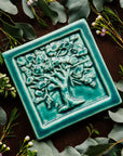 The Oak Tree Tile features a high relief oak tree, covered in layers of abstract leaves. The tile has a thick smooth border. This tile features the matte turquoise Pewabic Blue glaze.