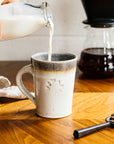 Cafe Mugs have a simple, modern design with smooth, straight sides that slant out. This makes the base slightly smaller than the cup's opening. The round Pewabic logo is stamped onto the side. This cafe mug features the creamy Birch glaze which has natural brown dappling and a grey interior glaze.