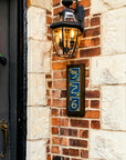 A three digit address frame is attached to a brick house below a porch light. The frame is made of a stained dark wood.