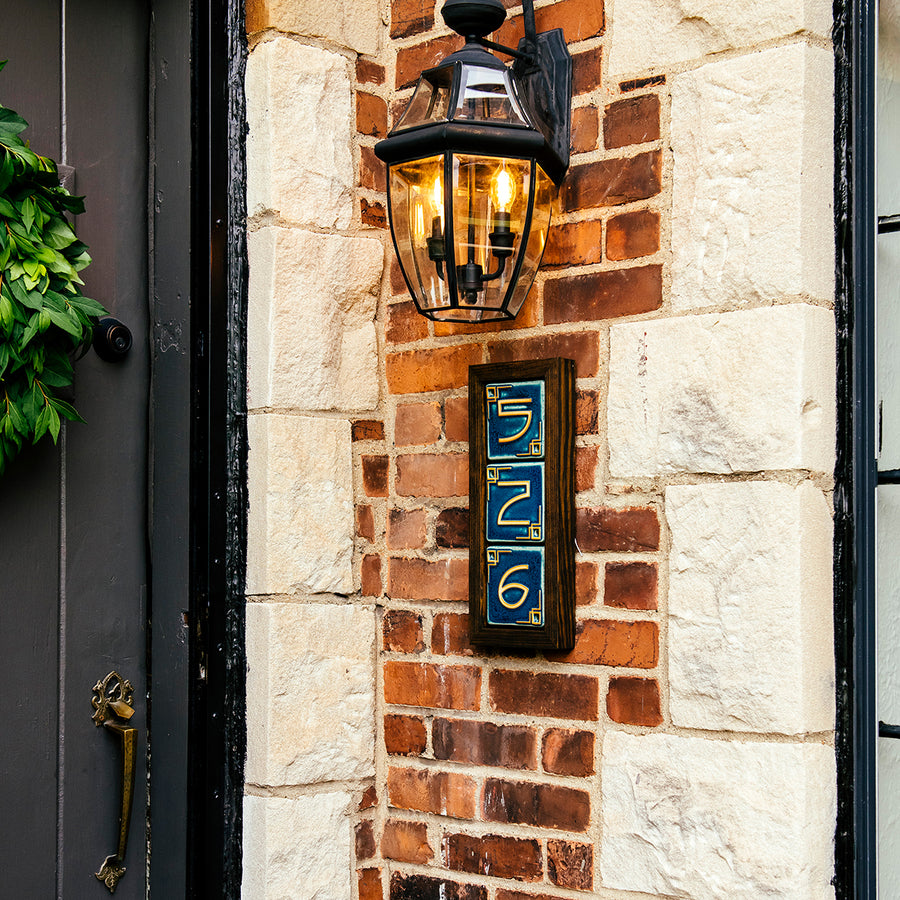 A three digit address frame is attached to a brick house below a porch light. The frame is made of a stained dark wood.